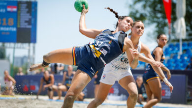 An athlete in a blue uniform leaps mid-air on a sandy court, holding a green ball, while another player in white shorts defends closely behind.