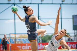An athlete in a dark blue uniform leaps to pass a green ball while being defended by an opponent in a white jersey.
