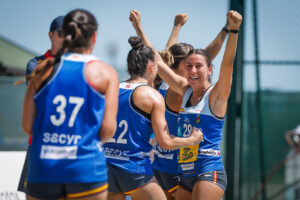 A group of athletes in blue jerseys celebrate enthusiastically with raised fists and joyful expressions on a sunny outdoor field.