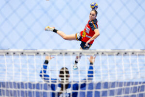 An athlete in a red and blue uniform kicks a handball mid-air during a game, with an opponent's raised hands visible in the foreground.