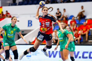 An athlete in a red and blue uniform leaps to pass a handball while being defended by players in green uniforms during a competitive match.