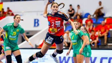 An athlete in a red and blue uniform leaps to pass a handball while being defended by players in green uniforms during a competitive match.