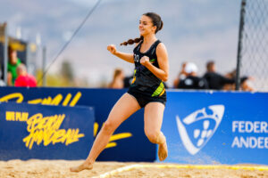An athlete in a black and green uniform leaps joyfully on a sandy court during a beach handball match, with spectators and branded banners visible in the background.